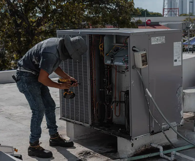 HVAC technician in a bucket hat servicing an open rooftop air conditioning condenser unit with a power drill
