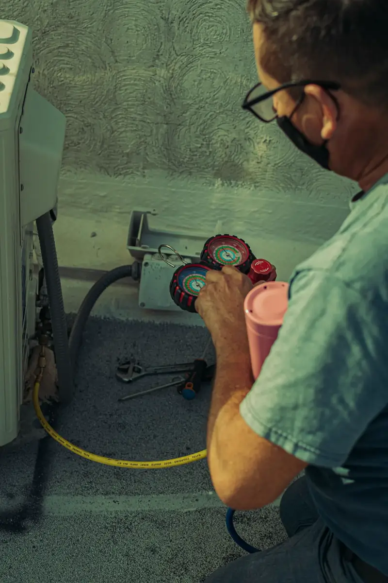 HVAC technician checking manifold gauges on an outdoor unit with tools laid out beside the equipment